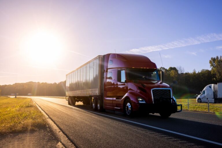 Red semi-truck driving on highway at sunset, surrounded by trees and clear blue sky.